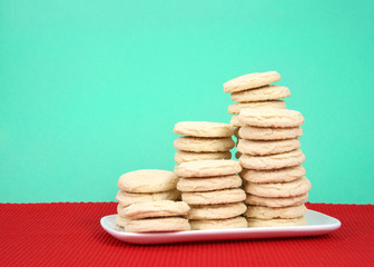 Sugar cookies stacked at multiple heights on a white rectangular plate red placemat, green background.