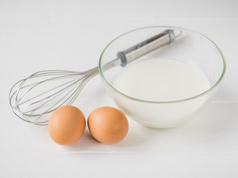 Two eggs, a whisk and a bowl of milk on white wooden table.