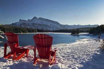 Red Adirondack Chairs and Distant Snowcapped Rocky Mountains Winter Landscape at Three Jack Lake in Banff National Park Alberta Canada - obrazy, fototapety, plakaty