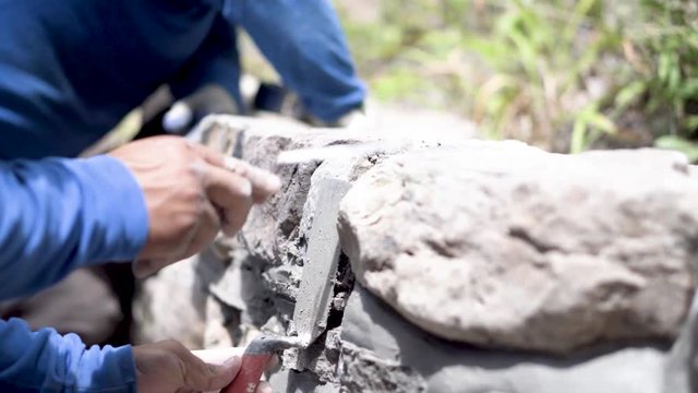 Holding a wide trowel as a barrier, a hardscape presses the mortar in between the rocks with a tuckpointing trowel.