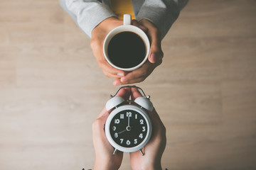 Women hands holding a cup of coffee and alarm clock showing it is wake up time in the morning
