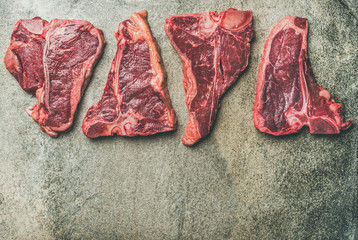 Flat-lay of fresh raw beef meat steak cuts over grey concrete background, top view, copy space. Porterhouse, t-bone and rib-eye steaks. Butcher' s shop or cooking meat dinner concept