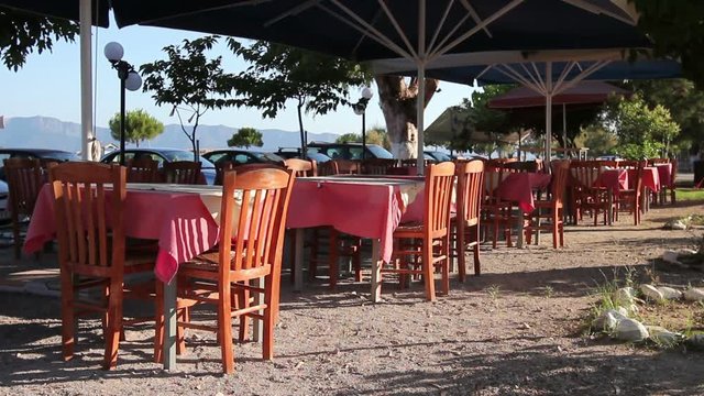 Chairs and tables in typical outdoor Greek tavern in morning sunlight with shadows on seashore.

