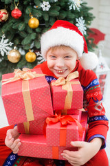 Closeup portait of smiling funny little kid holding many red presents decorated with golden ribbons. Small boy celebrating Christmas with family. Vertical color image.