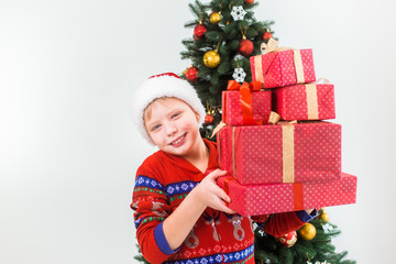 Obraz na płótnie Canvas Closeup portrait of happy smiling child holding pile of present boxes in hands ready to open them. Boy wearing santa hat and red pajamas. Holiday green tree in background. Horizontal color photography