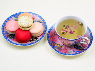 Cup of rose bud tea in a pretty floral cup and saucer served with a matching plate of traditional colorful French macarons over a white background