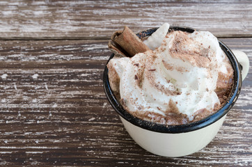 Enamel mug of hot chocolate cocoa with whipped cream and cinnamon stick on wooden table background. Winter times drink concept. Delicious cold weather beverage. Copy space.