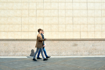 Side view portrait of two modern handsome men wearing autumn coats walking in city street against...