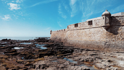 Fototapeta na wymiar Fortress of San Sebastian, low tide and stones covered with algae on a sunny day in Cadiz, Andalusia, Spain.
