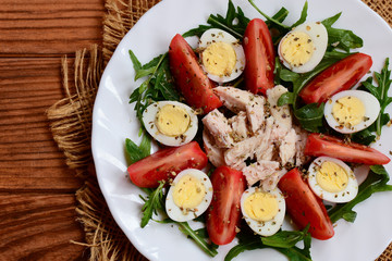 Vegetable salad with chicken breast and quail eggs. Salad with fresh tomatoes, rucola, quail eggs, boiled chicken breast and spices on a white plate and a wooden table. Closeup. Top view