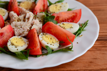 Homemade vegetable chicken salad for lunch or dinner. Healthy salad with fresh tomatoes, rocket, quail eggs, chicken fillet and spices on a white plate and a wooden background. Closeup