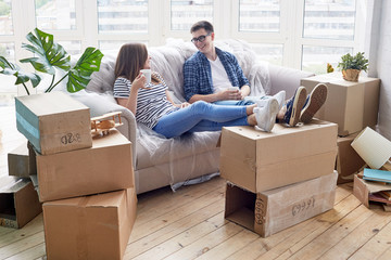 Cheerful young couple chatting animatedly with each other and drinking delicious cappuccino while taking short break from unpacking stuff, piles of moving boxes