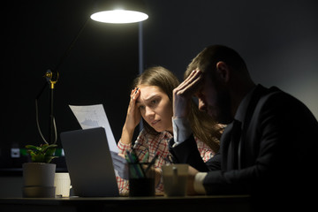 Two colleagues looking at bad company report in late evening meeting. Man and woman holding their...