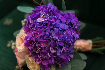Golden wedding rings on bouquet of purple hydrangea, pink roses flowers with lilac ribbons in rustic style.