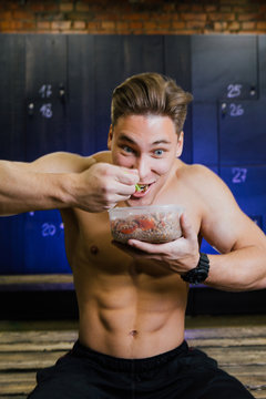 Fitness model of a bodybuilder guy eating food from a plastic container in a male locker room.