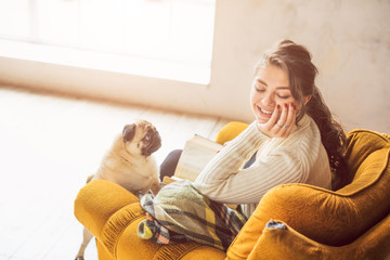 Smiling happy woman sitting on chair in sunny room holding book. Cute small pug pet want to play.