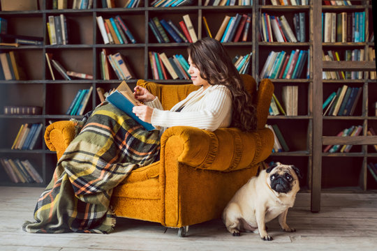 Reading time. Beautiful woman in cozy chair in library with her funny smart dog pug friend. Interesting literature world