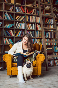 Young beautiful girl reading book sitting in the library. Funny dog pug sitting in her legs