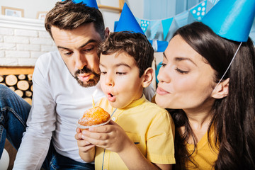 Let it come true. Cheerful lovely family blowing out a candle on a birthday muffin, making a wish while throwing a birthday party for their little son