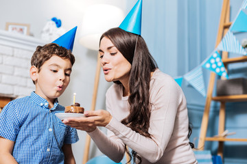 Tasty gift. Charming young woman holding a plate with a birthday cake and blowing out a candle on it together with her son while celebrating his birthday
