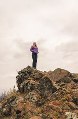 A girl in a lilac jacket looks out into the distance on a mountain, a view of the mountains and an autumnal forest by an overcast day, free space for text