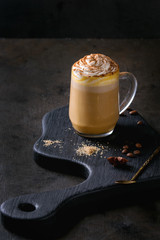 Glass of spicy pumpkin latte with whipped cream and cinnamon standing on black serving board. Coffee beans and spices above. Dark background.