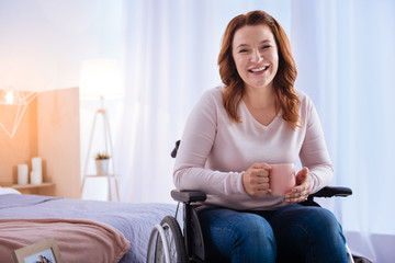 Happy moment. Exuberant blond disabled woman of middle age laughing and holding a cup while sitting...