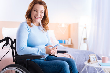 Texting friends. Attractive happy blond disabled woman of middle age smiling and using her phone while sitting in the wheelchair and a bed in the background