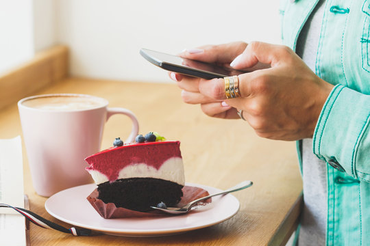 Woman takes a picture of cup with cappuccino and a piece of dessert on the plate