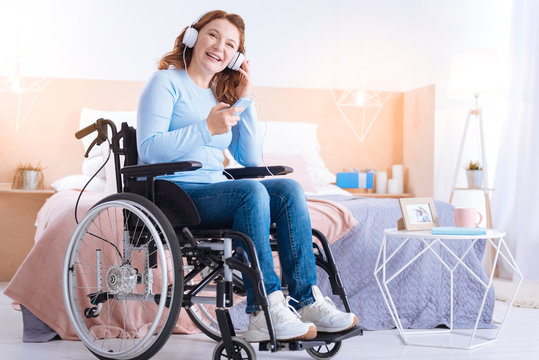 Time for music. Exuberant blond handicapped woman of middle age smiling and wearing headset and holding her phone while sitting in the wheelchair in a blue sweater