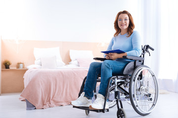 Fototapeta na wymiar I am satisfied. Smiling blond handicapped woman of middle age wearing glasses and holding a pen and a sheet of paper while sitting in a wheelchair in a blue sweater
