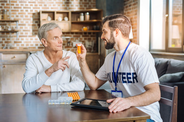 Useful vitamins. Young experienced cheerful volunteer looking friendly while showing new vitamins to an elderly man while visiting him