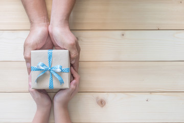 Father's day gift with daughter or son holding dad's hands giving present box with blue  ribbon on wood background to tell I love you dad and happy fathers day