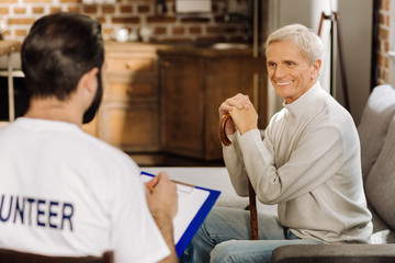 Obraz premium Cheerful pensioner. Pleasant positive elderly man looking happy while sitting near his kind attentive volunteer and smiling to him