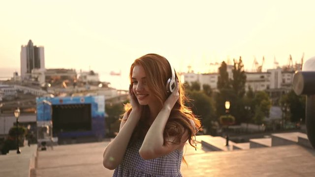Cheerful ginger lady with headphones looking camera while walking in the street 