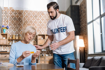 Feeling bad. Tired calm senior woman getting a pill from a little box while her attentive helpful social worker bringing her a glass of water