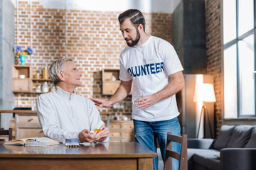 Bringing water. Helpful attentive young social worker giving a glass of water to a senior man sitting at the table and holding pills in his hands