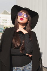 Glamorous young model wearing coat, broad brimmed hat and mirror glasses. Woman posing at the city in sun light