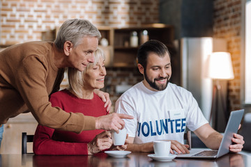 Amazing gadget. Cheerful clever senior people looking curious while a young pleasant enthusiastic volunteer showing them a modern laptop