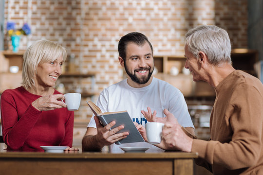 Exciting book. Positive young volunteer sitting at the table and holding an interesting book while talking to a pleasant aged couple