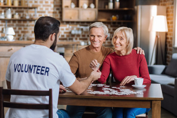 Lovely game. Cheerful elderly people hugging and looking at the young volunteer while sitting at the table and doing puzzles together