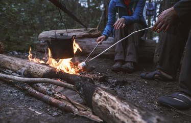 Two people are frying marshmelow and warming the kettle with water at the stake on a hike, on a warm summer evening.