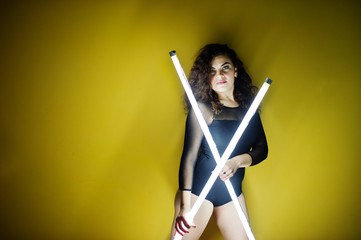 Curly brunette plus size model with two long led lamp against yellow wall.