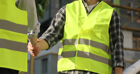Young caucasian builder in green uniform standing on unfinished construction territory, holding drawing and shaking hand of his collegue. Outdoor.