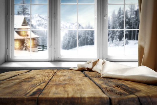 Wooden table with free space for your product or promotional text. Open window with snowfall. Landscape of winter forest mountains and small dock at the end of the village.