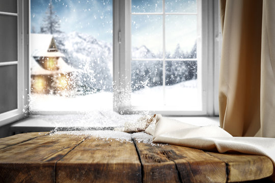 Wooden table with free space for your product or promotional text. Open window with snowfall. Landscape of winter forest mountains and small dock at the end of the village.