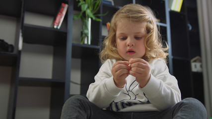 Caucasian pretty little girl with blond curly hair siting on the floor and opening the wrapper with toy on living background. Indoor.