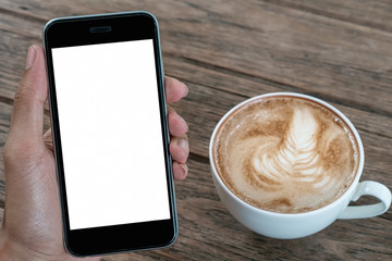 A man hand holding smart phone with latte hot, coffee cup on wood desk in coffee shop. mobile with blank screen and can be add your texts or others.