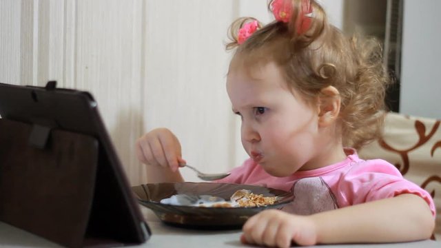 A little cute girl eats food and watches cartoons on the tablet. Child and gadgets, applications, internet, games.