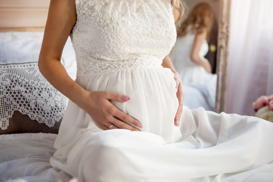 pregnant girl sits on the bed and holds on to the belly. She is dressed in a white long dress.